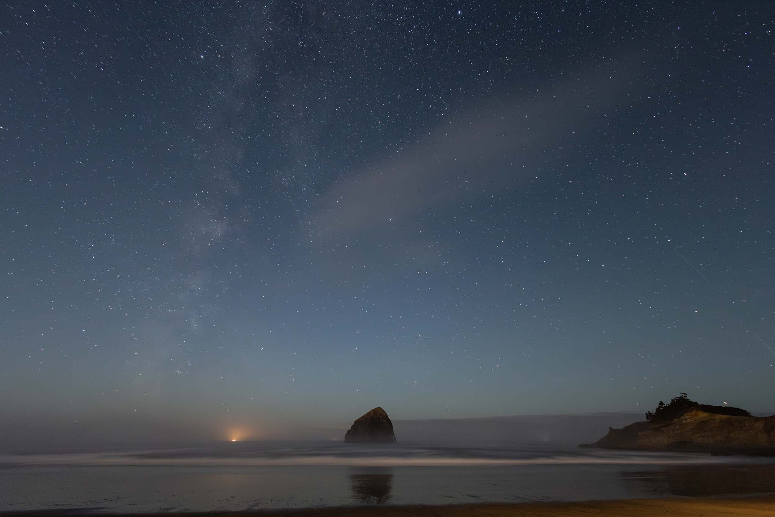 A view of Haystack Rock at night from the shore of the beach with a clear sky showing thousands of stars