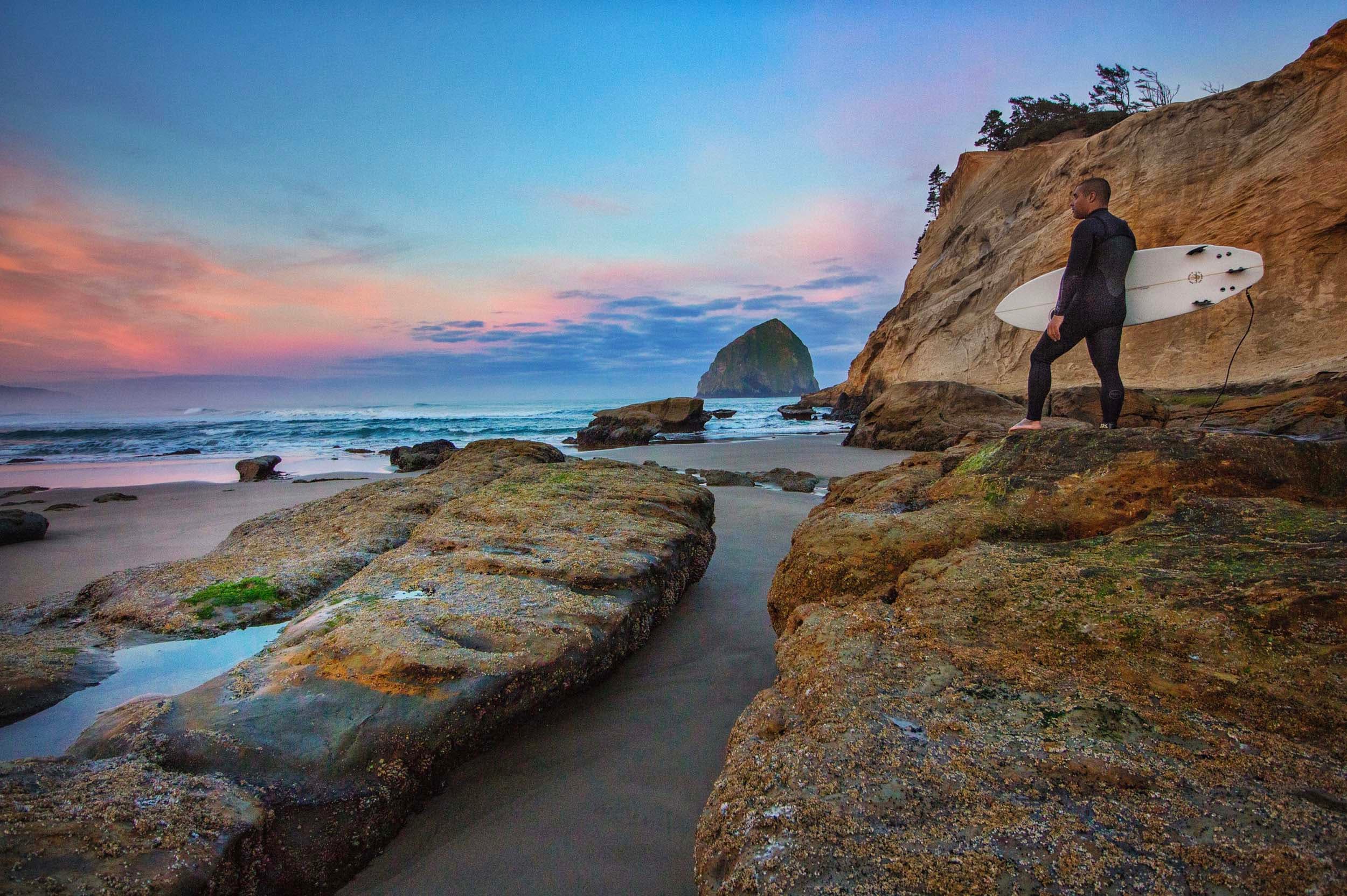 A man standing on a rock, holding his surfboard in the evening at Cape Kiwanda