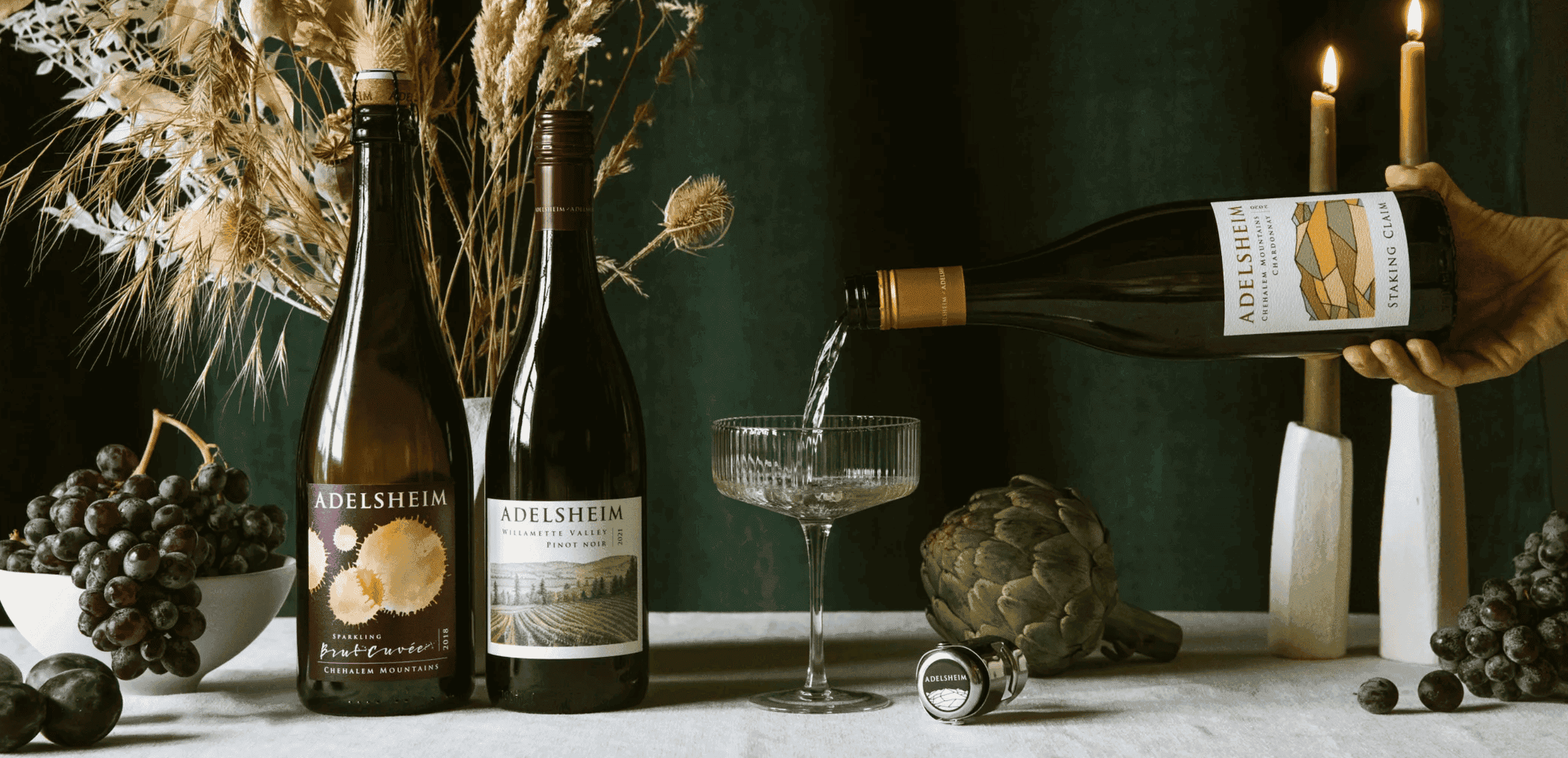 A hand pouring some chardonnay into a glass on a table with 2 other bottles of Adelsheim wine